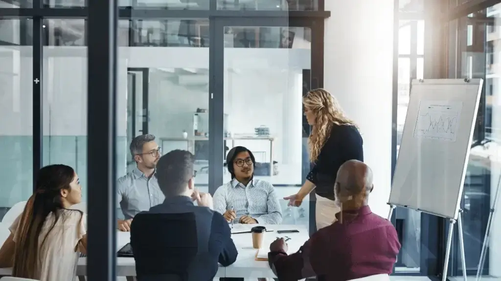 Imagem mostra uma equipe reunida em uma sala de um ambiente empresarial. Uma mulher está em pé explicando algo para a equipe.