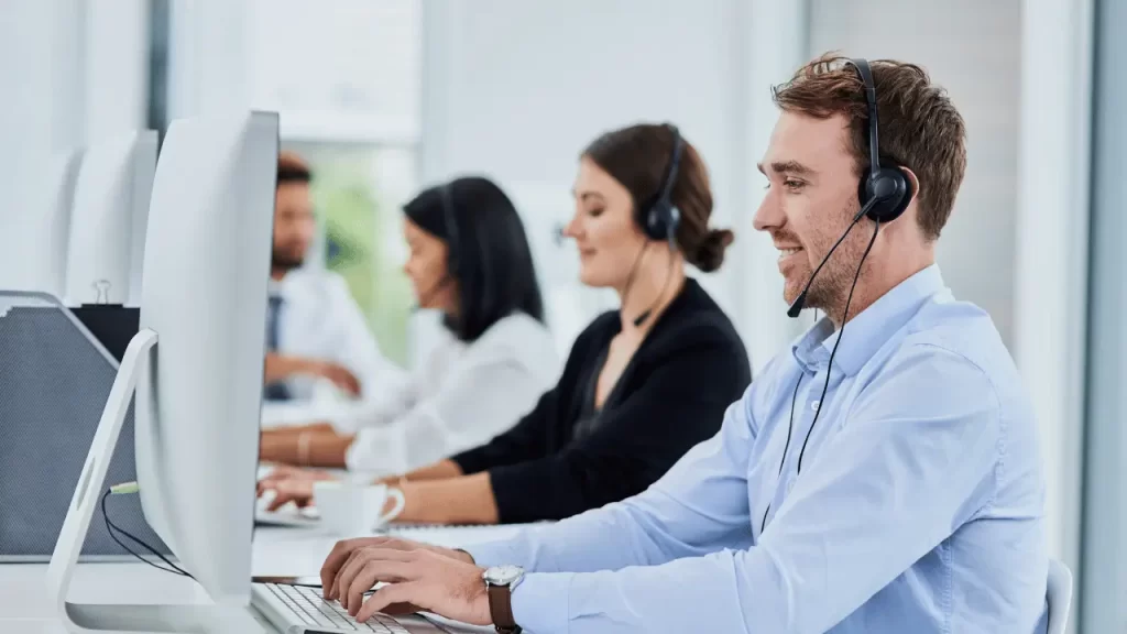 Pessoas em frente a computadores, com microfones no estilo tiara. São homens e mulheres trabalhando no atendimento.