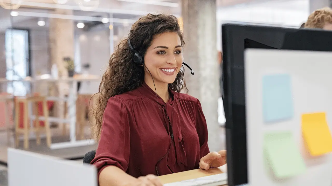 Mulher na frente de um computador, com um microfone tiara e camisa cor de vinho