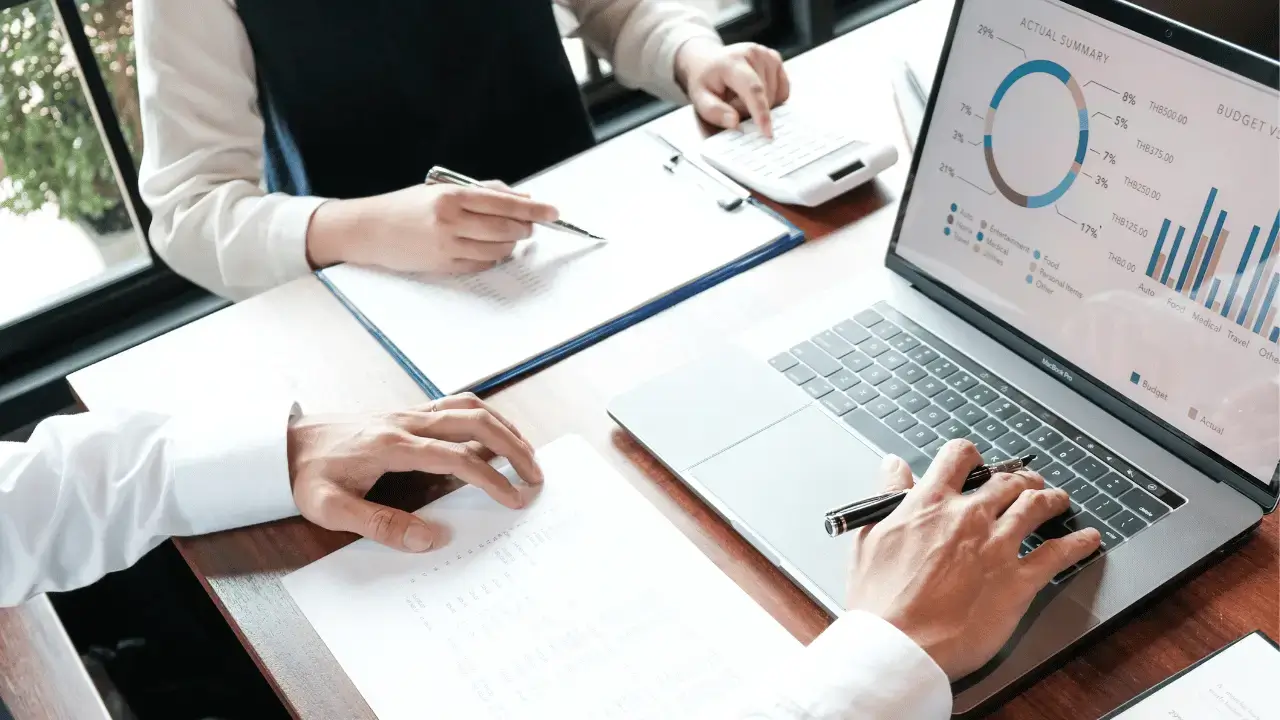 Dois homens analisando dados. Um deles na tela de um notebook e outro em um caderno. Os dois são brancos e vestem roupas sociais.