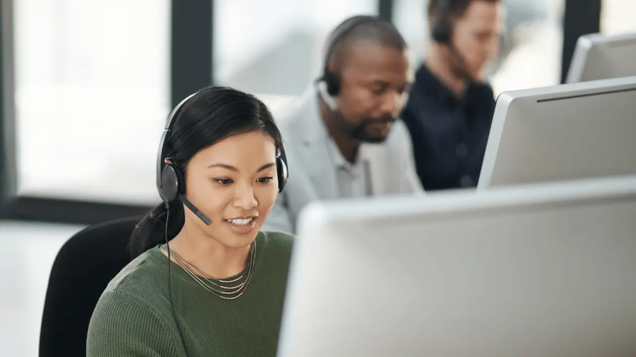 No primeiro plano está uma mulher com um fone tiara, estilo call center, olhando para a tela de um computador. Ela tem cabelos pretos e feições orientais. Atras está um homem negro com um microfone tiara também.