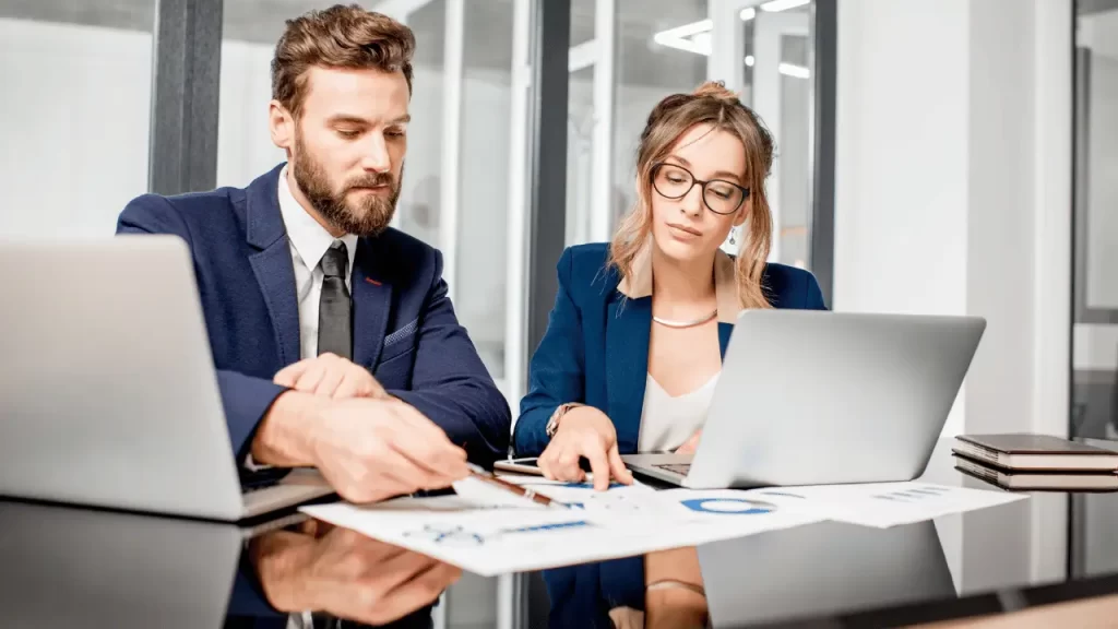 Homem e mulher analisando dados que estão em folhas em cima de uma mesa, com dois notebooks abertos ao lado. Homem e mulher são brancos e vestem roupas social.