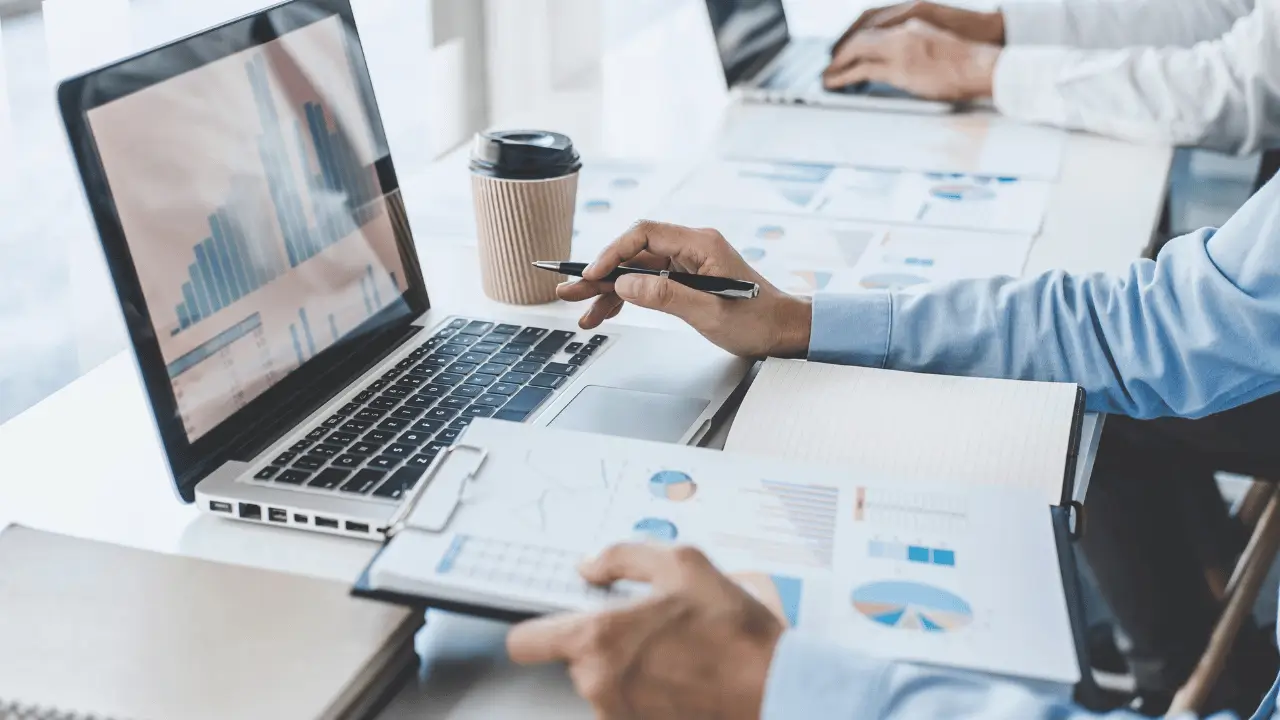 Homem branco, com camisa social, analisando dados na tela de um notebook e segurando documentos em papel