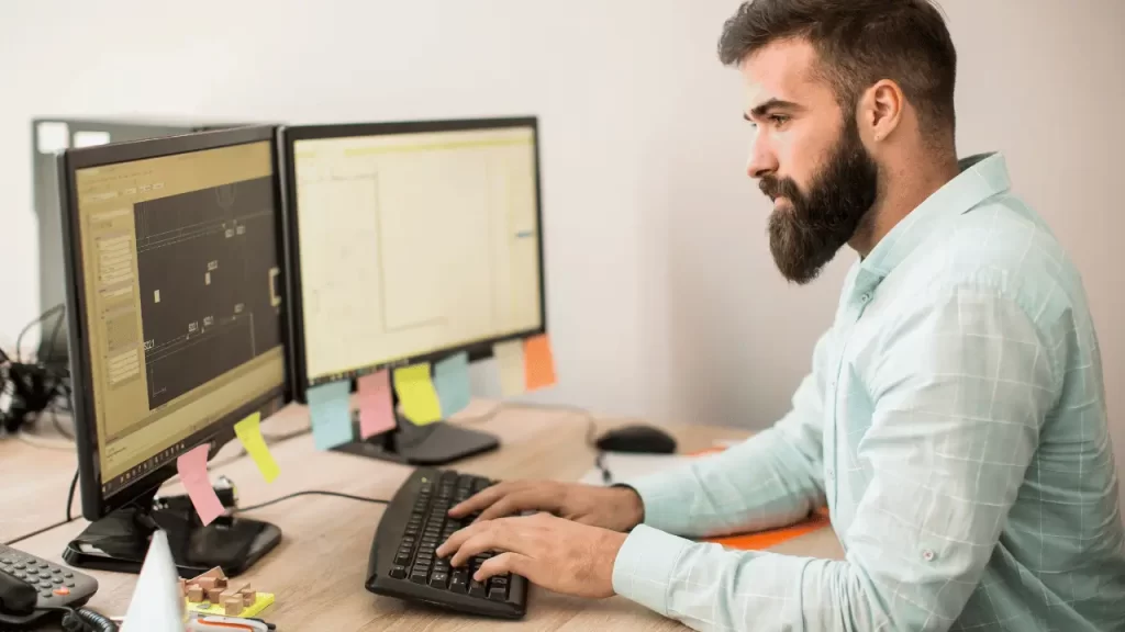 Homem trabalhando com duas telas de computador. Ele é branco, usa barba e veste camisa social azul.