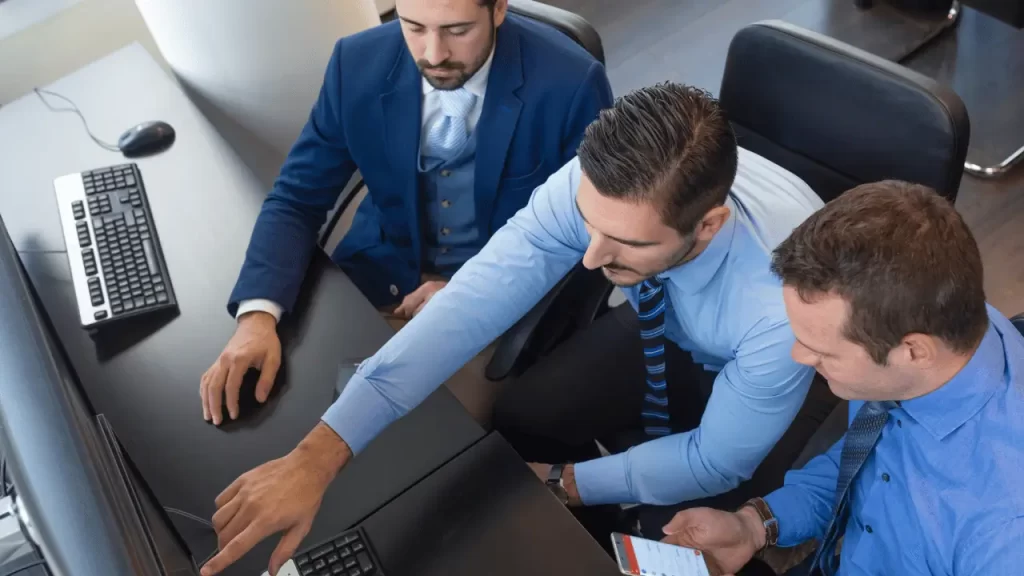 Três homens, todos brancos e vestindo roupas social,  analisando dados na tela de um computador, em um ambiente empresarial.