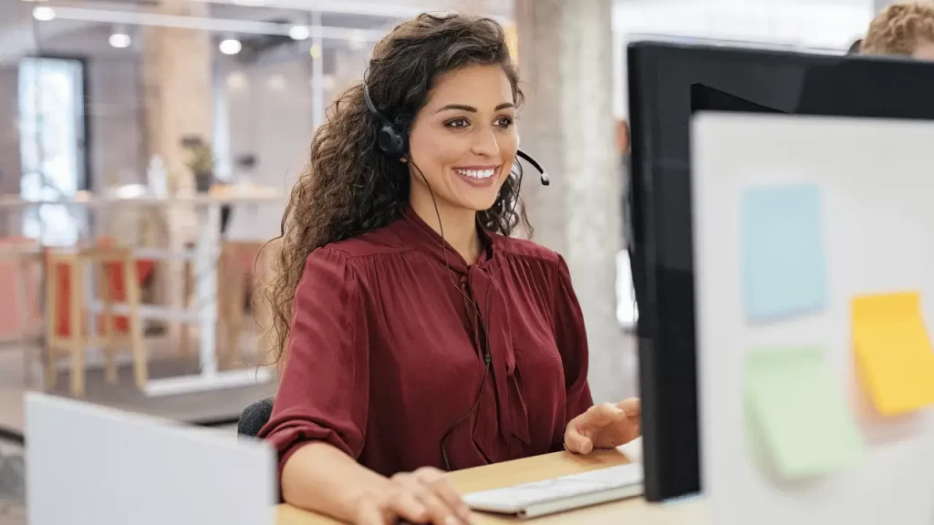 Mulher trabalhando de como call-center, com um microfone estilo tiara. Ela é branca e veste uma camisa social cor de vinho. 