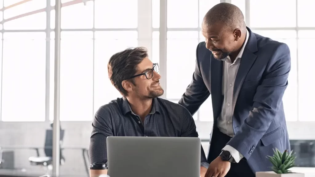 Cadeia de valor: o que é, 3 exemplos e como implementar Dois trabalhadores conversando em frente a um notebook.
